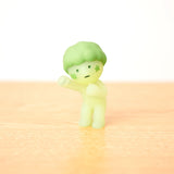 The Smiski Cheer Series - Blind Box by Smiski features a glow-in-the-dark figure with a green star on its cheek and hair, striking a fighting pose on a light wooden surface. Experience the excitement—each blind box holds a surprise.