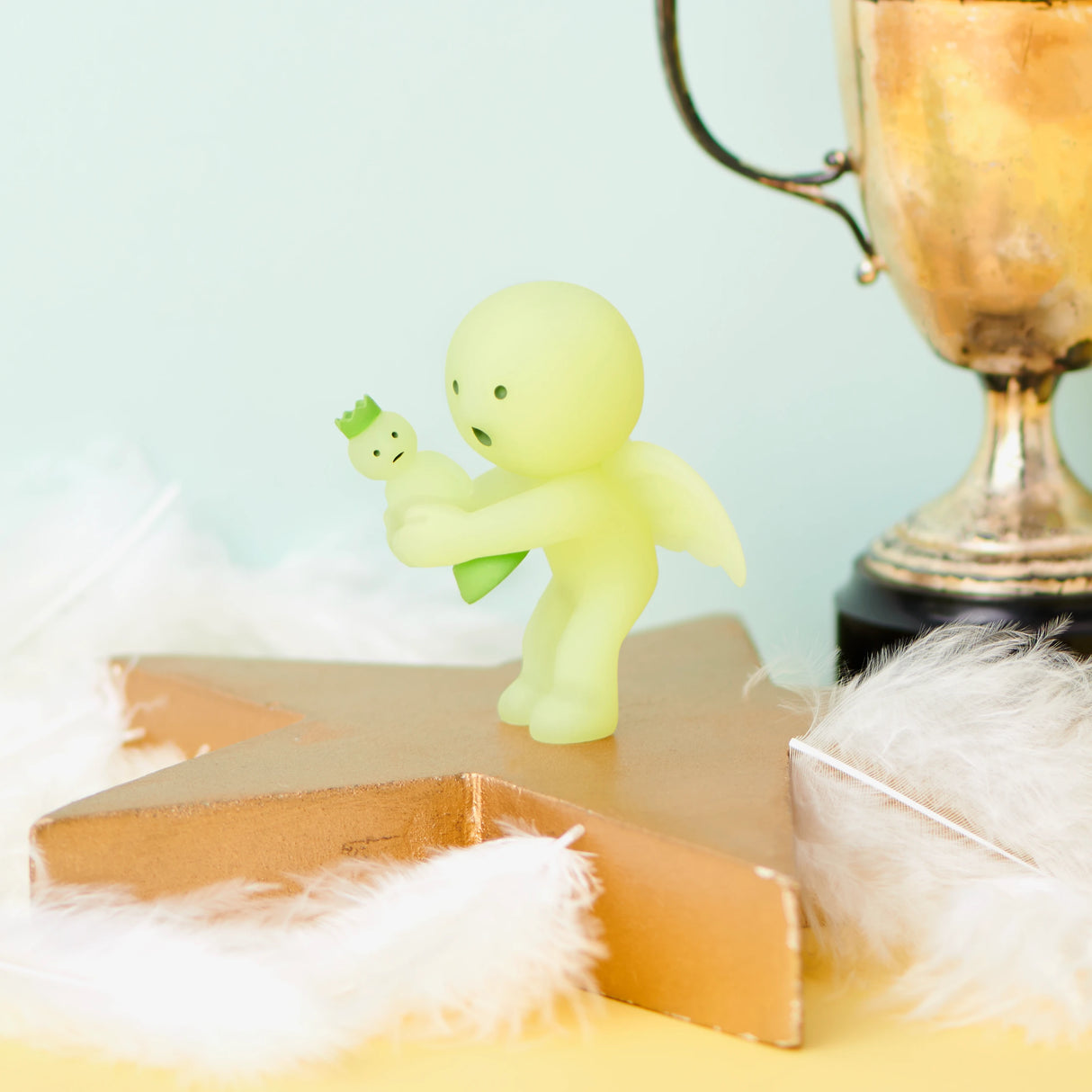 A small green figure from the Smiski Cheer Series - Blind Box, with outstretched arms, stands on a gold star platform holding a tiny companion. Nearby, white feathers and a gold trophy enhance the charm against a light blue background. It's a delightful addition to any Smiski collection.