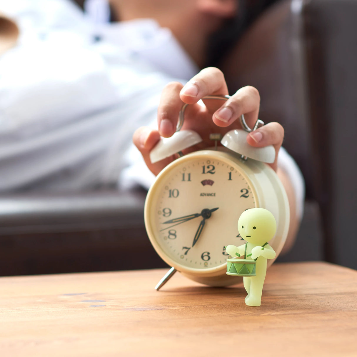 A sleepy person reaches for a ringing alarm clock next to a glow-in-the-dark figure from the Smiski Cheer Series - Blind Box by Smiski, resting on a wooden surface.