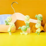 Five small figures from the Smiski Dressing Series - Blind Box, in green and yellow glow-in-the-dark colors, are posed in various positions against a yellow background, with a clothes hanger and a striped cloth nearby.