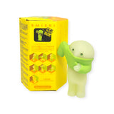 A small figurine wearing a green shirt and glowing in the dark is positioned next to a yellow box labeled "Smiski Dressing Series - Blind Box," which showcases illustrations of similar characters from the series.