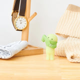 A small figurine from the Smiski Dressing Series - Blind Box, adorned with a scarf, stands on a wooden surface alongside a watch on a stand, a striped shirt, and a knitted sweater. Its glow-in-the-dark charm adds an enchanting touch to the display.