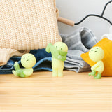 Three Smiski Dressing Series figures, featuring glow-in-the-dark, green, round heads and small bodies, are arranged on a wooden surface in front of folded clothes. These playful figurines from the Smiski brand bring charm to any room as you look forward to your next Smiski Dressing Series - Blind Box surprise.