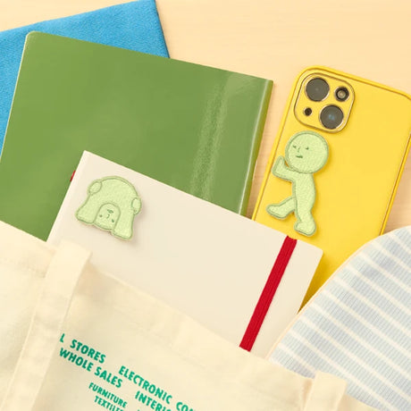 Three light green Smiski embroidery sticker patches in new poses on notebook, phone, and tote bag handle on wooden surface
