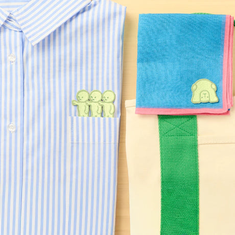 Three pale green Smiski embroidery sticker patches peeking out of shirt pocket and one on folded blue cloth near beige tote bag