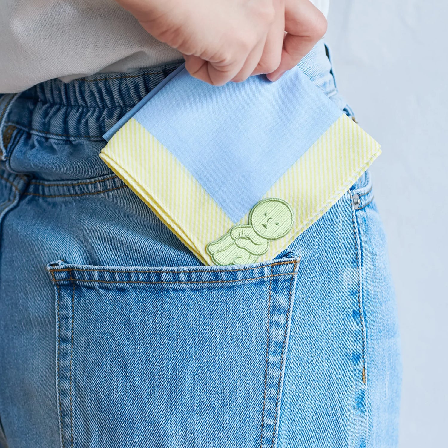 Someone tucks a folded blue and yellow handkerchief with a small Smiski Embroidery Sticker/Patch by Smiski into the back pocket of blue jeans.
