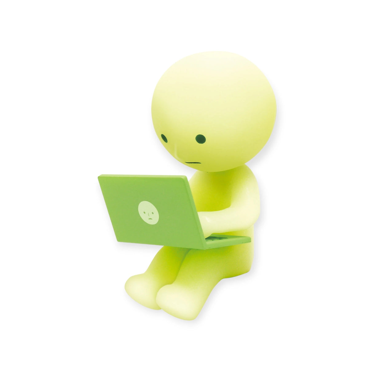 Seated with a green laptop, the small, green cartoon figure from the Smiski At Work Series - Blind Box by Smiski showcases its round head and minimal facial features. This charming character possesses a delightful glow-in-the-dark feature.