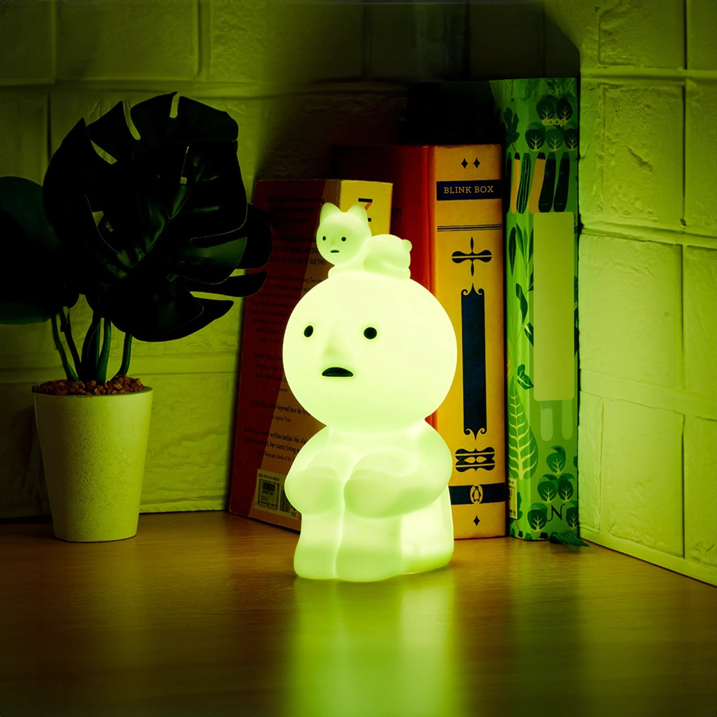 The Smiski Touch Light by Smiski, a delightful glowing lamp featuring an adorable cartoon figure with a tiny cat on its head, rests on the table as a nightlight surrounded by books and a potted plant. This touch-activated whimsical piece brings warmth and charm to your reading nook.