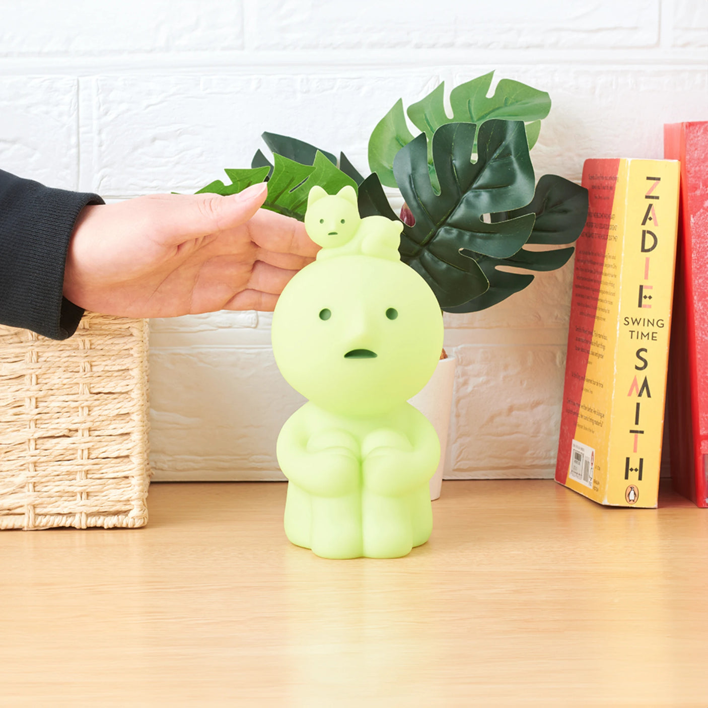 A person's hand reaches for the Smiski Touch Light, a quirky green lamp designed like a cartoon character featuring a smaller figure on its head. This delightful touch-activated nightlight sits on a wooden surface alongside books and a plant.