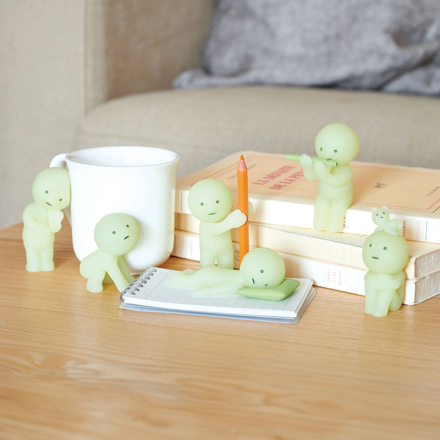 Five small, green humanoid figures from the Smiski Living Series - Blind Box by Smiski are arranged around a white mug, books, and a notepad with a pencil on a wooden table. Their subtle glow-in-the-dark feature adds an unexpected twist to your collection.