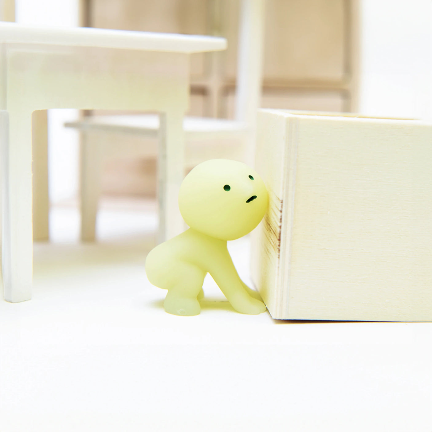 A Smiski Living Series - Blind Box figure, from the Smiski brand, takes the intriguing shape of a small, round, green character leaning on a wooden block amidst white furniture. This glow-in-the-dark collectible adds a mysterious flair to your space, reminiscent of the surprise elements characteristic of a blind box reveal.