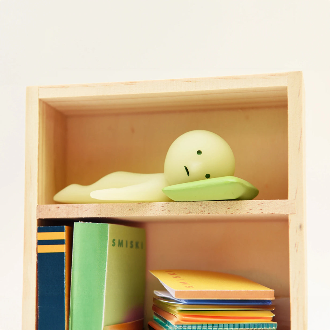 A small green figure from the Smiski Living Series - Blind Box by Smiski lies on its side atop the wooden bookcase, glowing in the dark above a stack of multicolored notebooks.