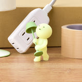 A Smiski Moving Series - Blind Box collectible toy, holding a green umbrella, is positioned beside an electrical outlet and a roll of tape on a wooden surface. This glow-in-the-dark figure from Smiski brings a whimsical touch to any environment.