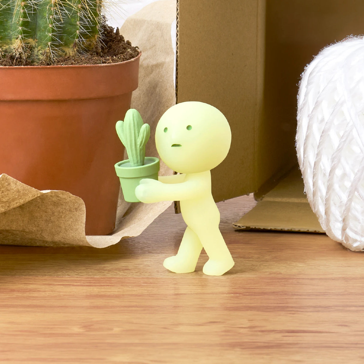 A small figurine from the Smiski Moving Series - Blind Box, presented in yellow, gently holds a tiny green potted cactus next to a larger cactus plant and some white twine on a wooden surface. This delightful collectible toy by Smiski is perfect for adding character to any space.