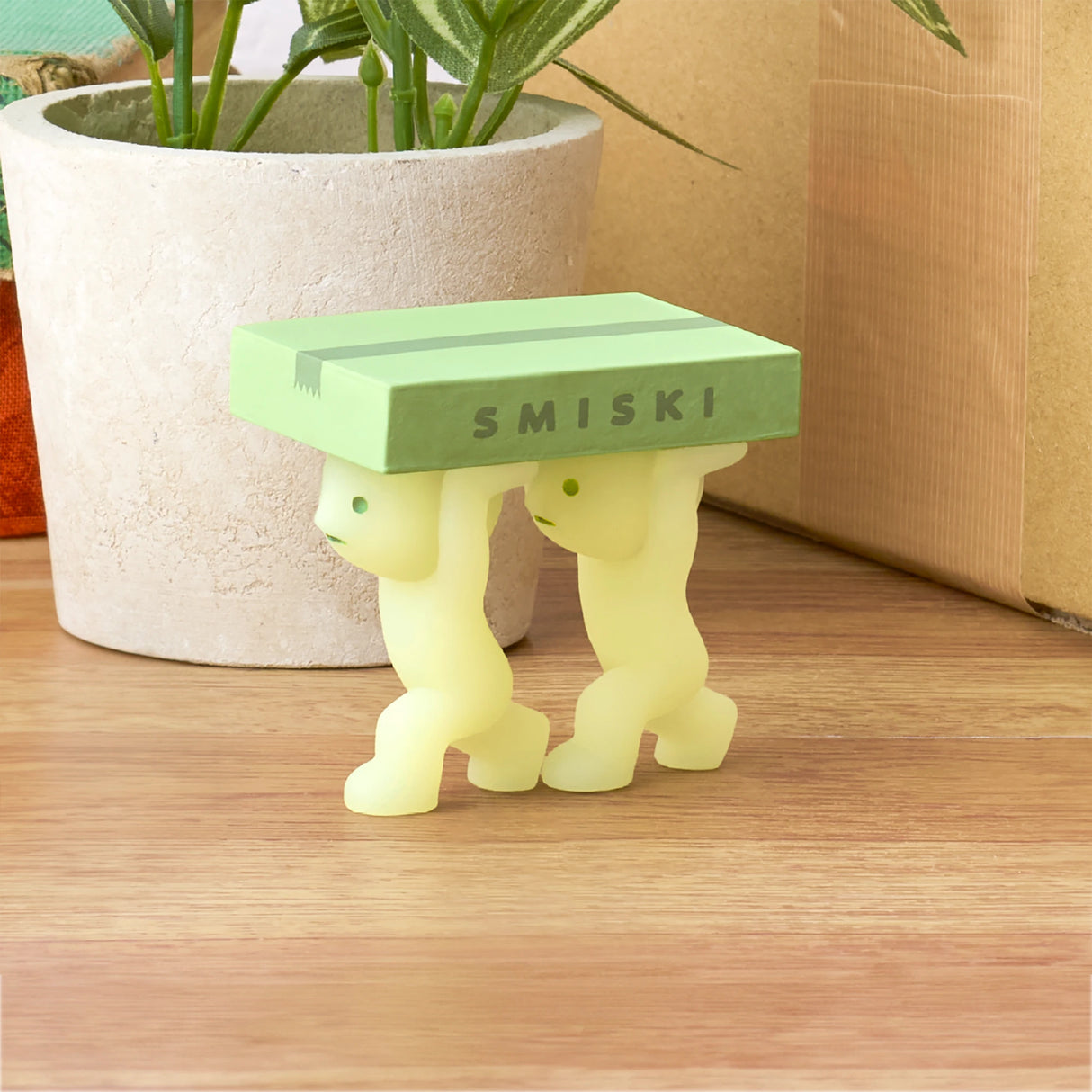 Two small figurines from the Smiski Moving Series by Smiski hold up a green rectangular block together on a wooden surface near a potted plant. As charming collectible toys, they add a whimsical touch to any space with their glow-in-the-dark feature.