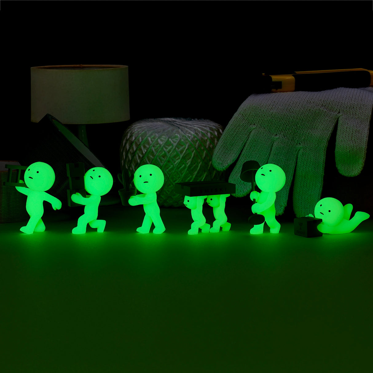 Six small figures from the Smiski Moving Series - Blind Box, each glow-in-the-dark humanoid striking different poses, are arranged on a table. In the background, a lamp, yarn, glove, and tape measure add to this whimsical scene of collectible toys.