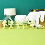 The Smiski Moving Series - Blind Box by Smiski features small green figurines arranged in a row, each holding items such as boxes and tape. Behind them, a surface adorned with a lamp, cutting tool, twine, and glove adds to the scene. These glow-in-the-dark collectible toys bring a whimsical touch to any setting.