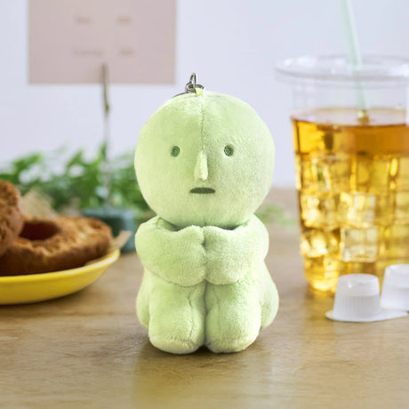 A small, light green Smiski Plush Keychain - Hugging Knees from Smiski sits upright on a table next to an iced drink, donuts, and coffee creamer cups.