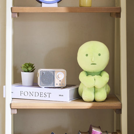 Light green Smiski Plush figure sitting with arms crossed around legs on wooden shelf near retro radio and small plant