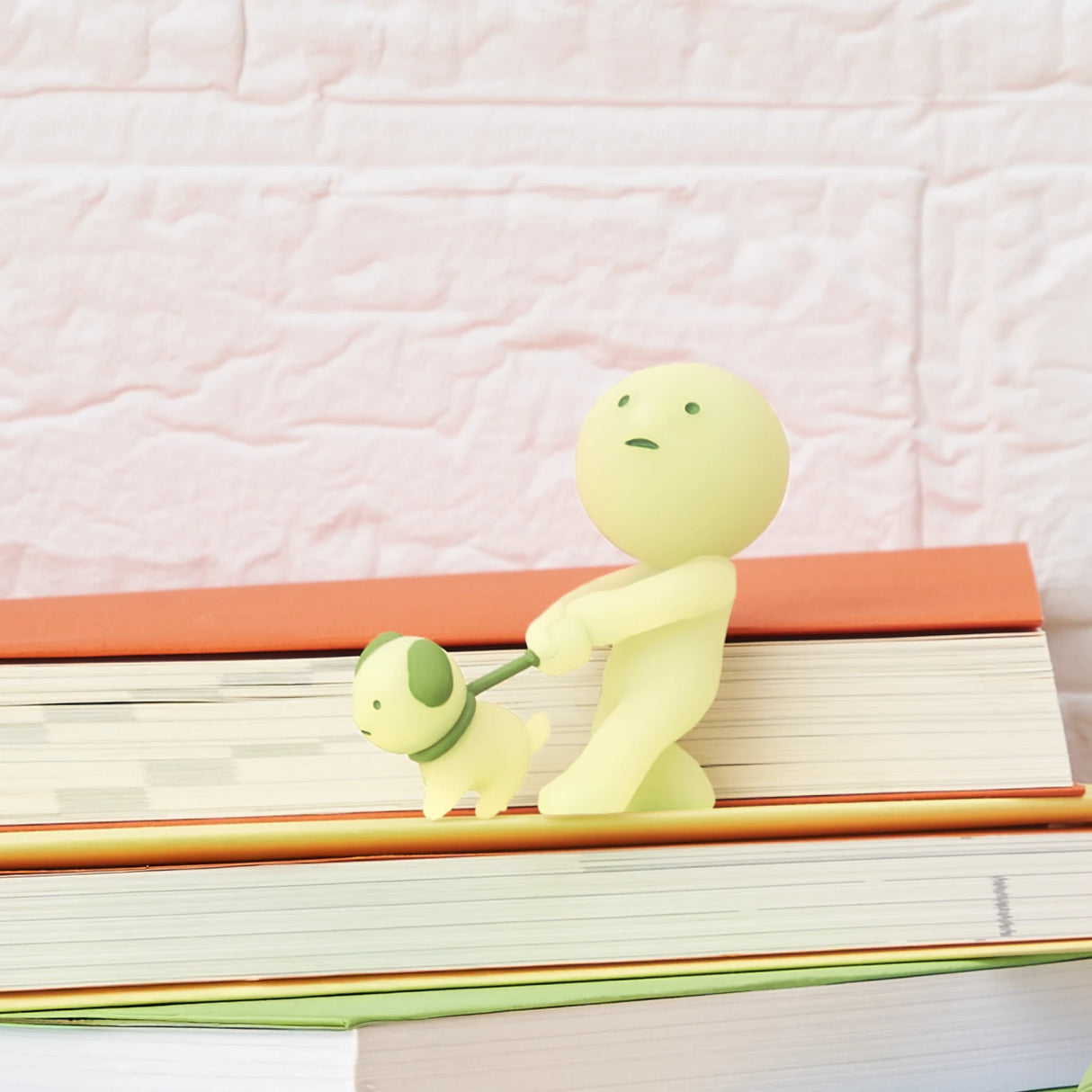 The Smiski Sunday Series - Blind Box by Smiski features a small, pale yellow figurine of a person walking a dog on books with a brick-patterned background—ideal for collectors and glow-in-the-dark enthusiasts.