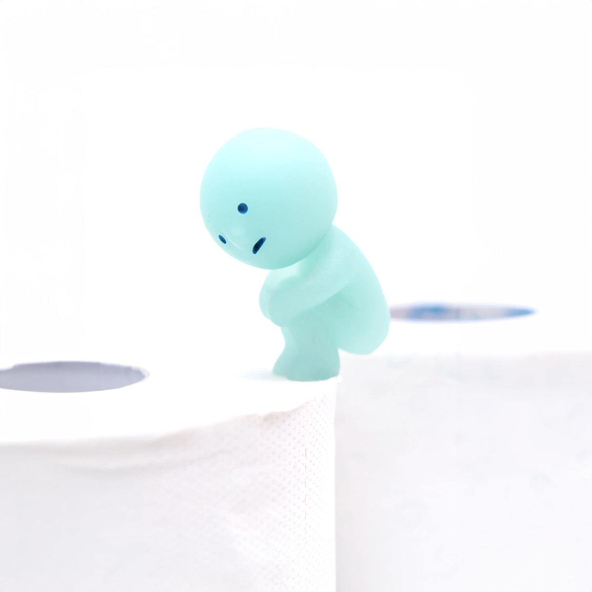 A small figure from the Smiski Toilet Series - Blind Box by Smiski stands cautiously on the edge of a toilet paper roll, seemingly contemplating a leap to an adjacent roll while highlighting its glow-in-the-dark allure.