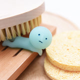 A small figurine from the Smiski Toilet Series - Blind Box by Smiski, featuring a neutral expression and oval shape in blue, is positioned next to a wooden brush and two round sponges.