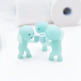 A trio of diminutive, light green glow-in-the-dark figures from the Smiski Toilet Series - Blind Box by Smiski hold tiny toilet paper rolls, positioned on a white surface with larger rolls in the background. Ideal for fans of whimsical mini collectible figures.