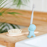 A Smiski Toothbrush Holder - Carrying Figure from the brand Smiski sits on a wooden counter, its phosphorescent glow-in-the-dark material adding a whimsical touch. A toothbrush is inserted, and a hairbrush rests beside it.