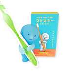 A small blue figure holding a green and white toothbrush is positioned beside a box labeled "Smiski Toothbrush Holder - Holding Figure," which features an illustration of the same figure. This delightful Smiski product from the Smiski brand adds a whimsical touch and glow-in-the-dark charm to your bathroom decor while keeping it organized.