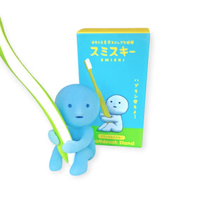 The Smiski Toothbrush Holder - Protecting Figure, a glow-in-the-dark figurine gripping a large toothbrush, stands proudly next to a matching blue box decorated with Japanese text and the words "Toothbrush Stand" in English, ready to be your protective pal.