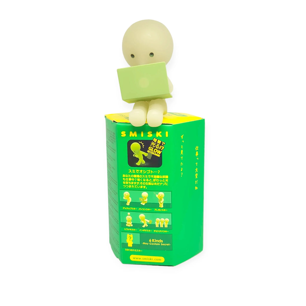 A small, green glow-in-the-dark Smiski figure sits on a box with a laptop. The Smiski At Work Series - Blind Box showcases various poses, making it an intriguing item to explore.