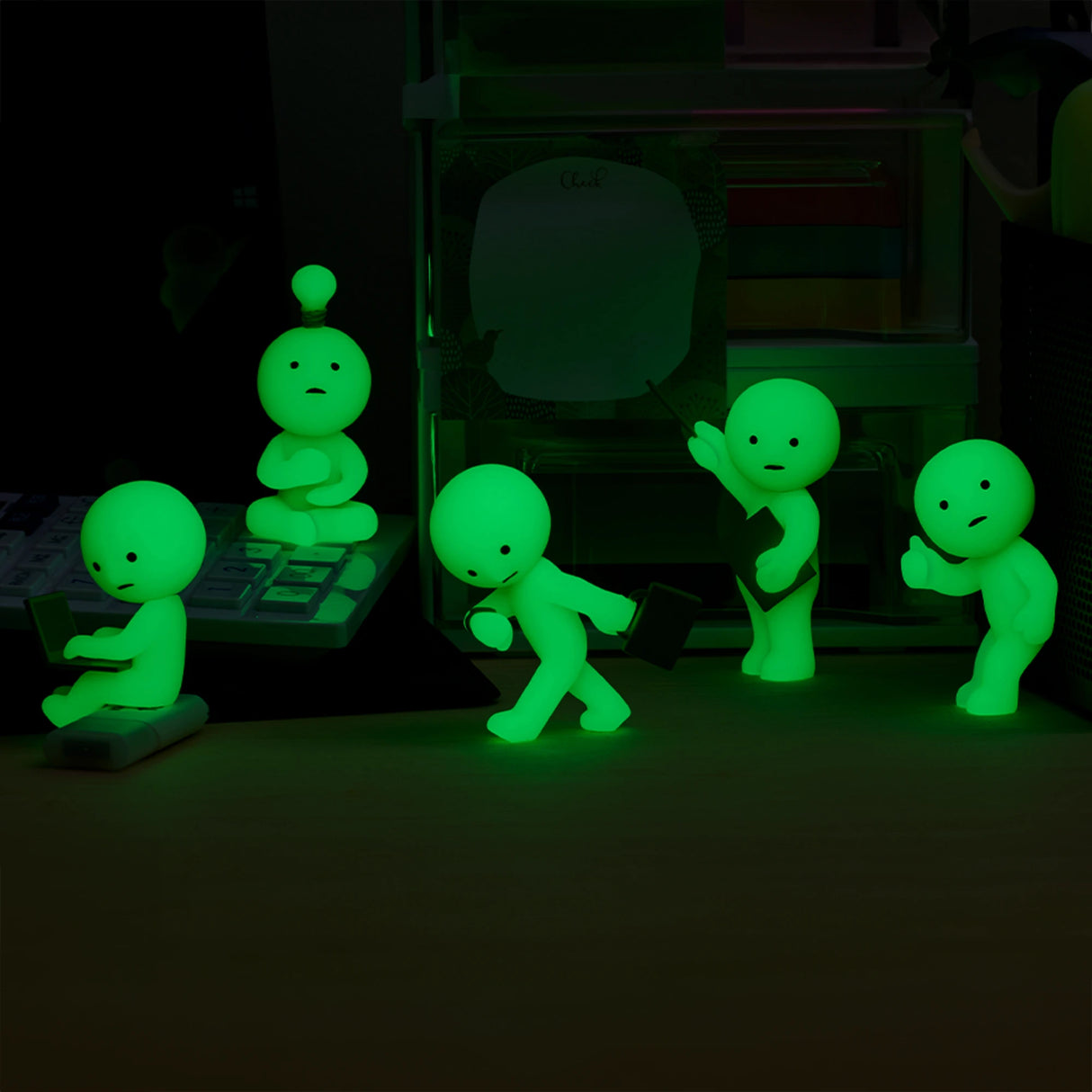 Five figurines from the Smiski At Work Series by Smiski, each in a unique green glow-in-the-dark pose, are placed on a desk.