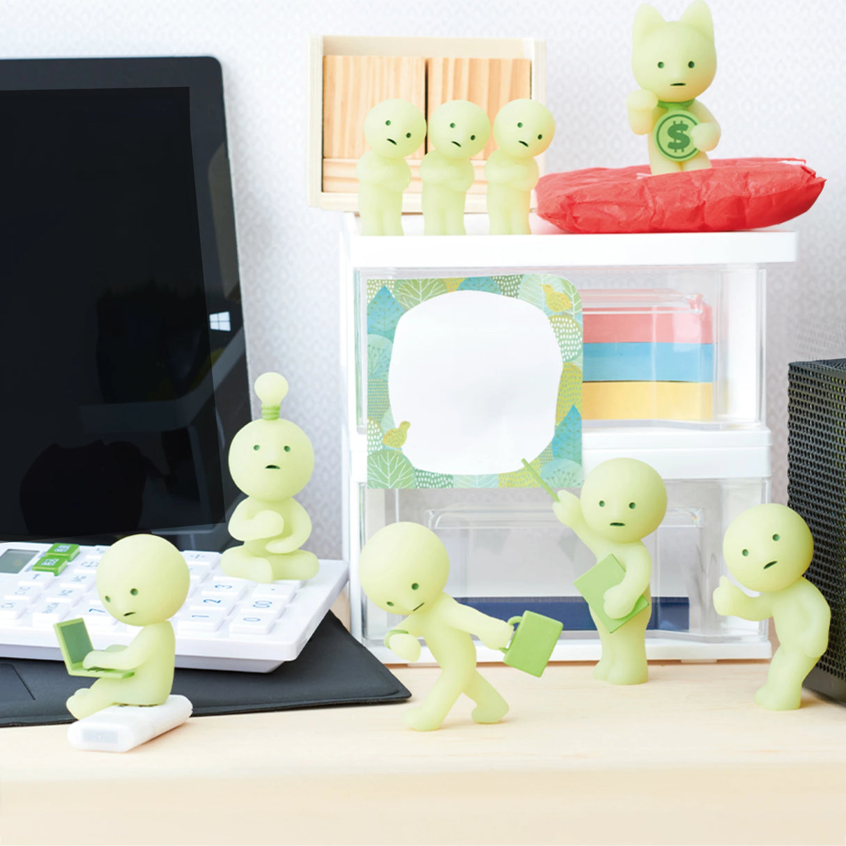 Small figurines from the Smiski At Work Series - Blind Box by Smiski are scattered around a desk featuring a calculator, computer, and organizers. These green characters seem to interact with office items and each other, bringing a whimsical element to your workspace.