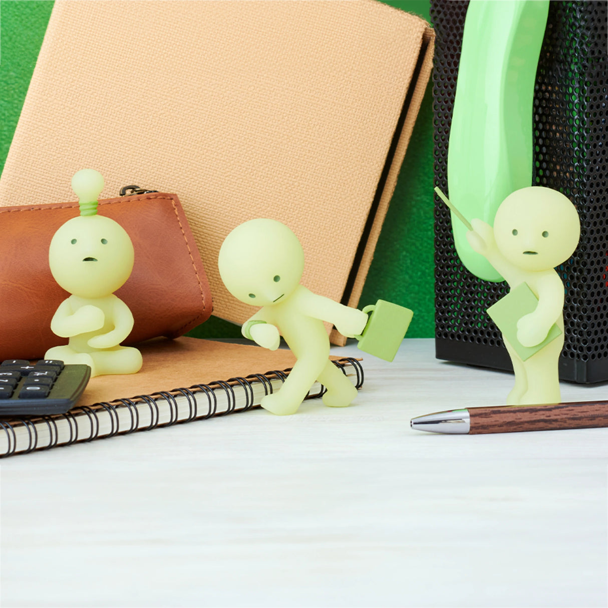 Three small figurines from the Smiski At Work Series by Smiski, in light green and playful poses, adorn a desk with notebooks, a calculator, and a pen. These delightful blind box collectibles even glow in the dark, bringing charm to any workspace.