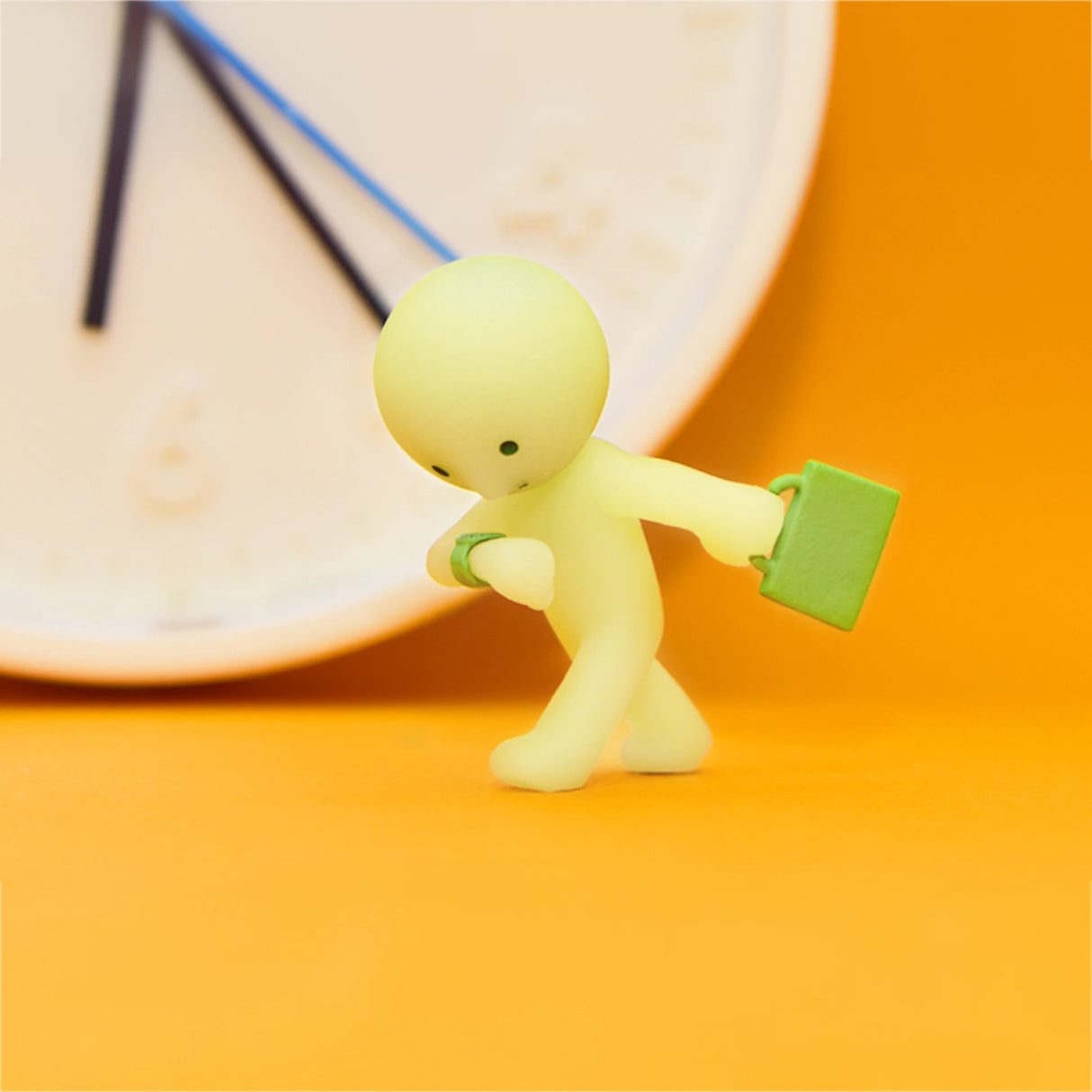 A tiny green figure from the Smiski At Work Series - Blind Box by Smiski holds a suitcase and glances at its watch, poised for its next journey. In the background, a large wall clock stands blurred against an orange surface, subtly suggesting the glow-in-the-dark secret tucked within this blind box gem.