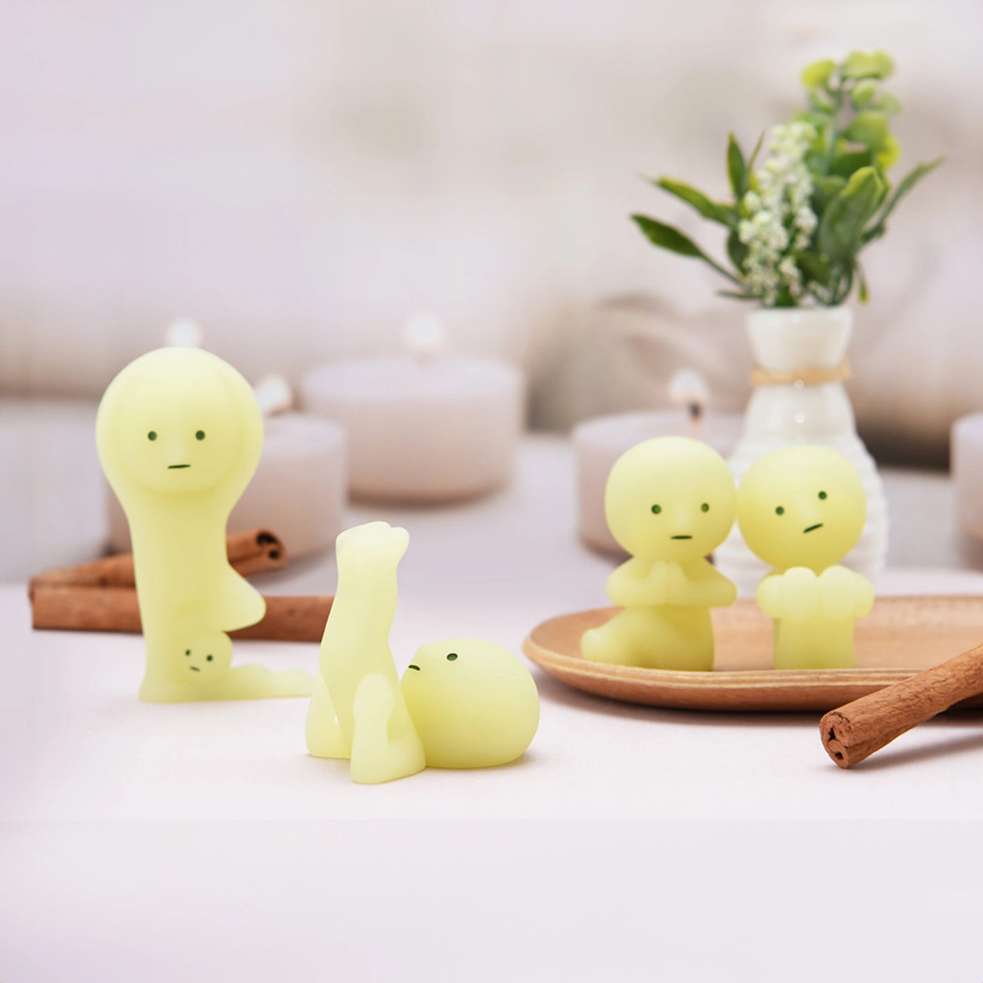 Four figures from the Smiski Yoga Series, each small with a pale green hue and simple facial features, are displayed on a table alongside cinnamon sticks and a vase of flowers. These glow-in-the-dark figurines by Smiski capture calming yoga poses, radiating an aura of tranquility.