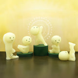 Against a yellow background, five small figurines from the Smiski brand grace the scene, each with glow-in-the-dark features and dark green bases. They belong to the Smiski Yoga Series - Blind Box and capture different tranquil yoga poses.