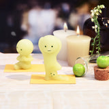 Two adorable figurines from the Smiski Yoga Series - Blind Box by Smiski, each featuring cute expressions and calming yoga poses, are placed on yellow mats next to candles and decorative items. Together, they create a playful scene on a flat surface, while their whimsical glow in the dark adds an enchanting touch.