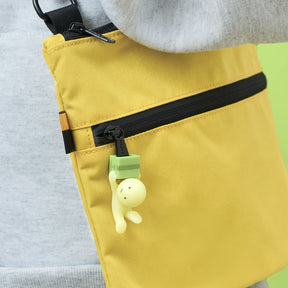 Close-up of a yellow bag with a black zipper, featuring a Smiski ZipperBite - Falling Down glow-in-the-dark figure keychain by Smiski attached to the zipper pull.