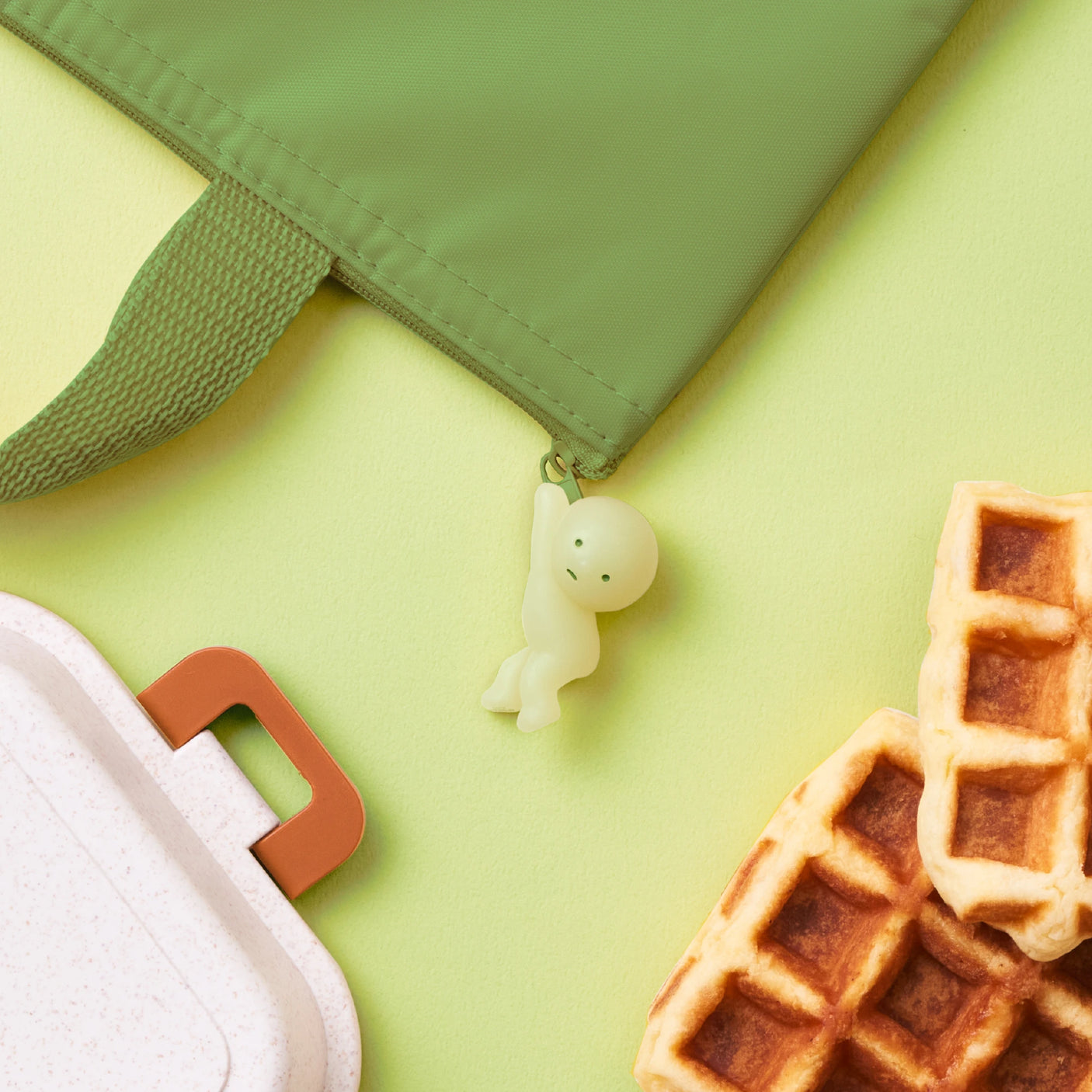 A Smiski ZipperBite - Hanging On figure by Smiski hangs from a green bag against a matching green background, glowing in the dark beside tempting waffles and the side of a white container with an orange handle.