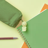 A green spiral notebook and orange paper overlap on a light surface. Nearby, a green pouch featuring a small Smiski ZipperBite - Hugging Knees glow-in-the-dark charm is partially open. A wooden pen rests beside them, subtly highlighting the charming zipper accessory from Smiski.