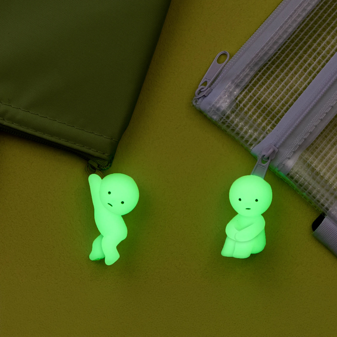 Two glowing green Smiski ZipperBite figures from the Smiski brand are featured on different bags; one appears to be climbing while the other is in a seated position. These playful accessories, named "Smiski ZipperBite - Hugging Knees," bring a whimsical touch to your belongings. They glow in the dark, adding both cuteness and functionality.