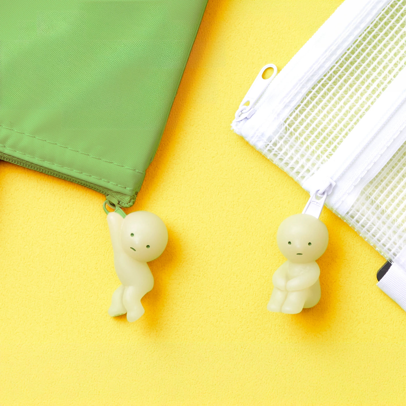 Two Smiski ZipperBite - Hanging On figurines from Smiski are secured to the zipper pulls of green and white pouches. The bright yellow background highlights the charming glow-in-the-dark appeal of these adorable zipper figures.