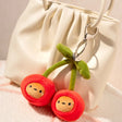 Plush cherry bag charm with metal keyring on white bag; green stem and red faces