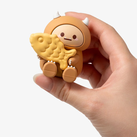 Hand holding a small brown Smoko Hank Yeti figure clutching a yellow fish-shaped street food snack on white background