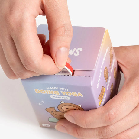 Hands opening a pastel blue and purple Smoko Hank Yeti Doing Yoga blind box with cartoon figure illustrations visible