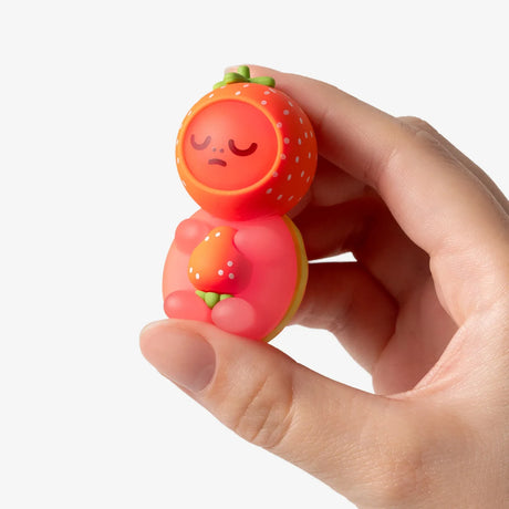 Hand holding a small sleepy Smoko Kawawa Turtle figure dressed in a strawberry costume against white background