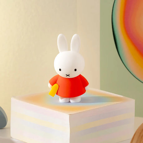 White bunny figurine in red dress holding a yellow object standing on multicolored pastel block with abstract background