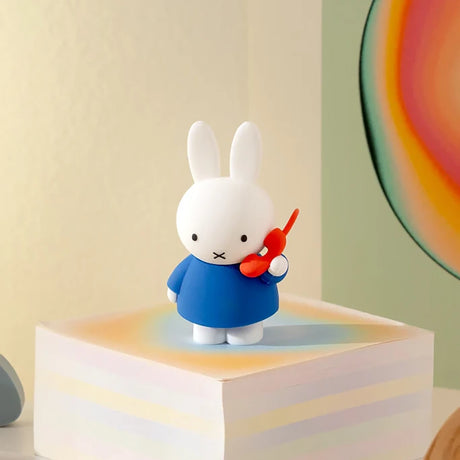 Small white bunny figurine in blue dress holding a red phone standing on a pastel-colored block