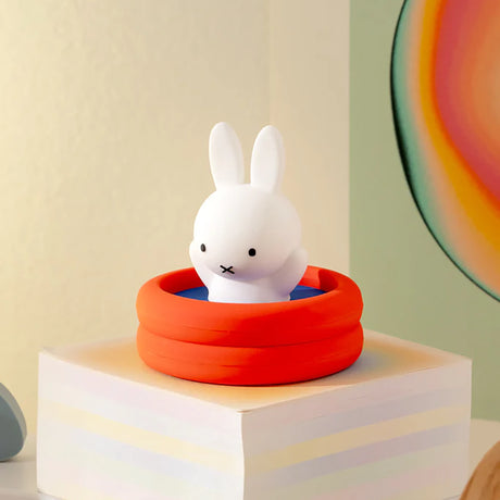White bunny figurine with a neutral expression sitting inside a small bright orange inflatable pool on a pastel block